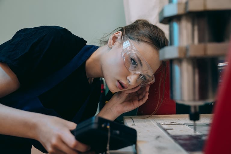 Young woman in workshop wearing goggles, focusing on machinery operation.